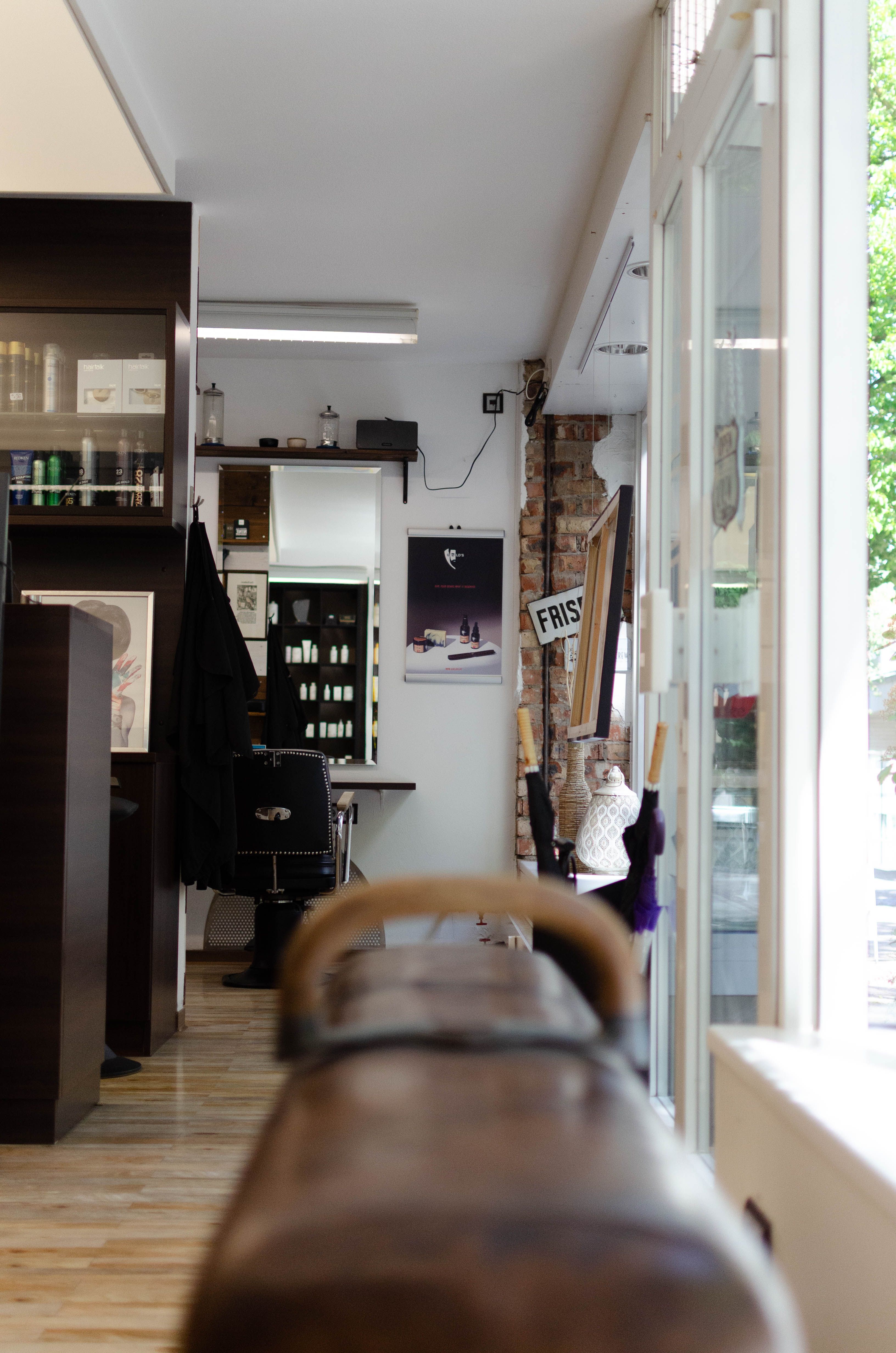 Blick über das Turnpferd an der Eingangstür und der Rezeption vorbei in den Herrenbereich mit klassischen Barber-Stühlen und Spiegeln, einer Backsteinwand und einem Schild das 'Friseur' schreibt